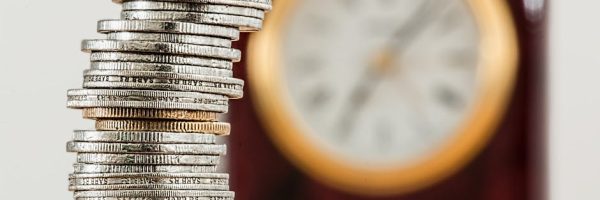 A close-up image of stacked coins with a blurred clock, symbolizing time and money relationship.